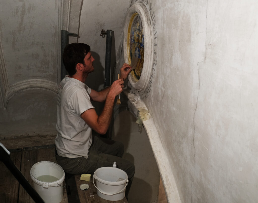 Notkonservierung Wand-/Deckenbilder, Dreibeinskreuzkapelle, Bürenstrasse 54, Solothurn. Die Malschicht wird mit Methylcellulose (3% in Wasser) konsolidiert. Das Konsolidierungsmittel wird über Japanpapier mit einem Pinsel aufgebracht.